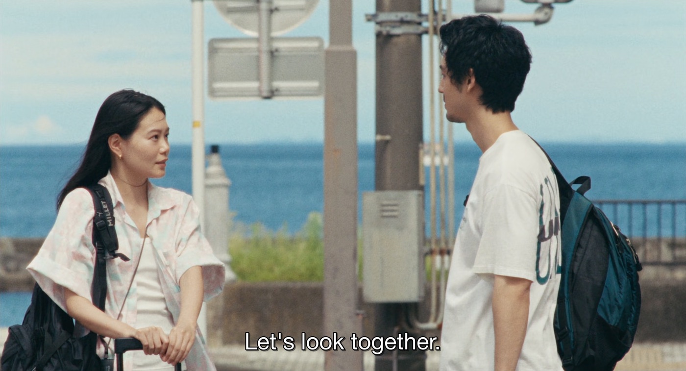 Film still of two people talking by the sea, subtitle reads "Let's look together."