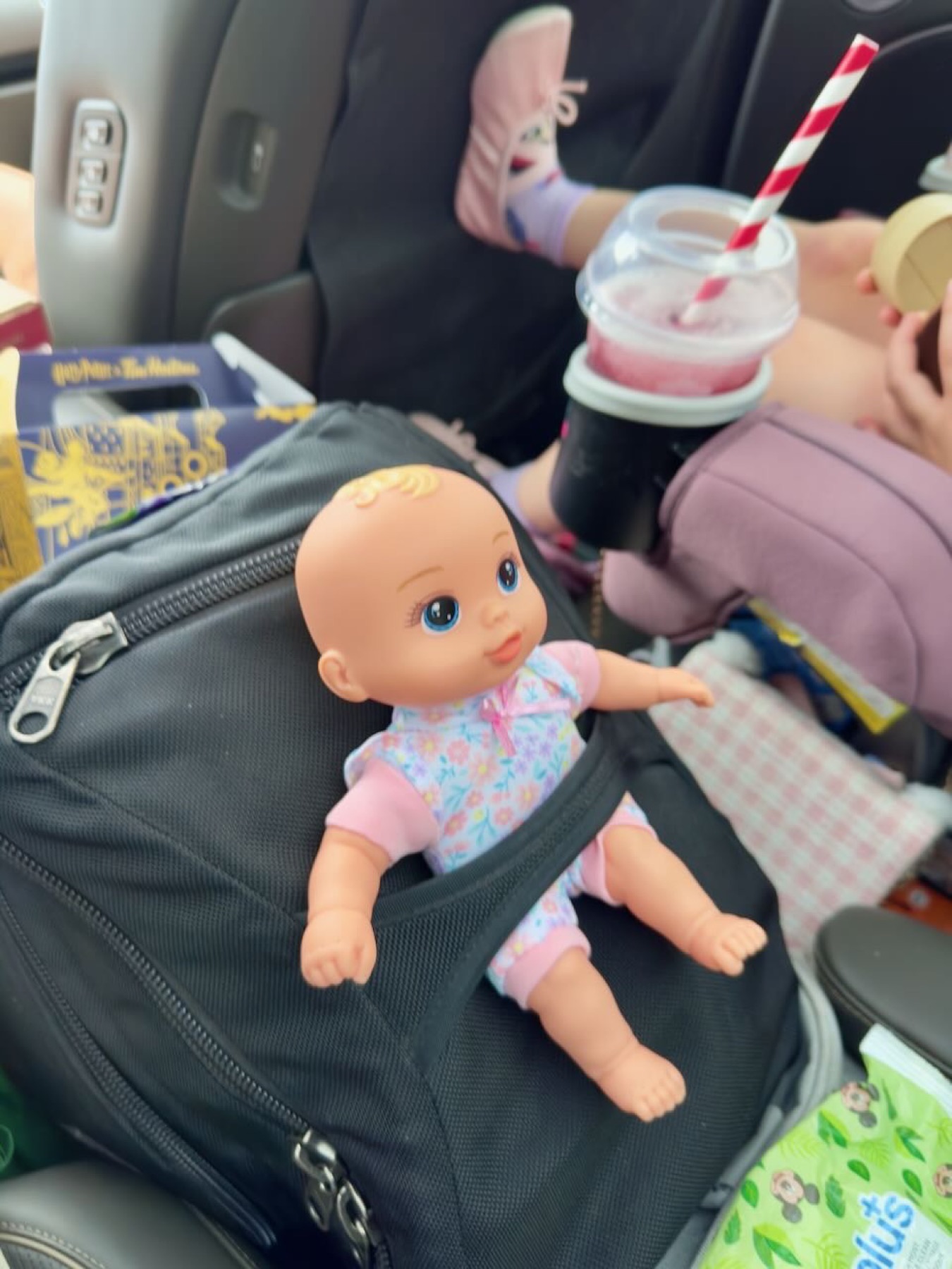 A doll propped on a backpack in the back of the car, slushie in the cup holder