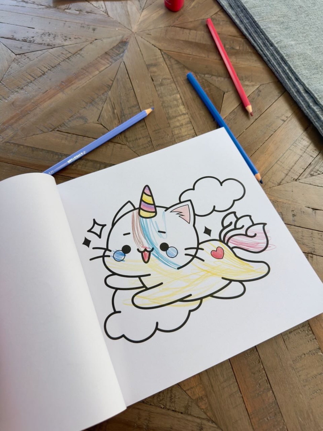 A unicorn cat part-coloured in a colouring book, pencils on the table